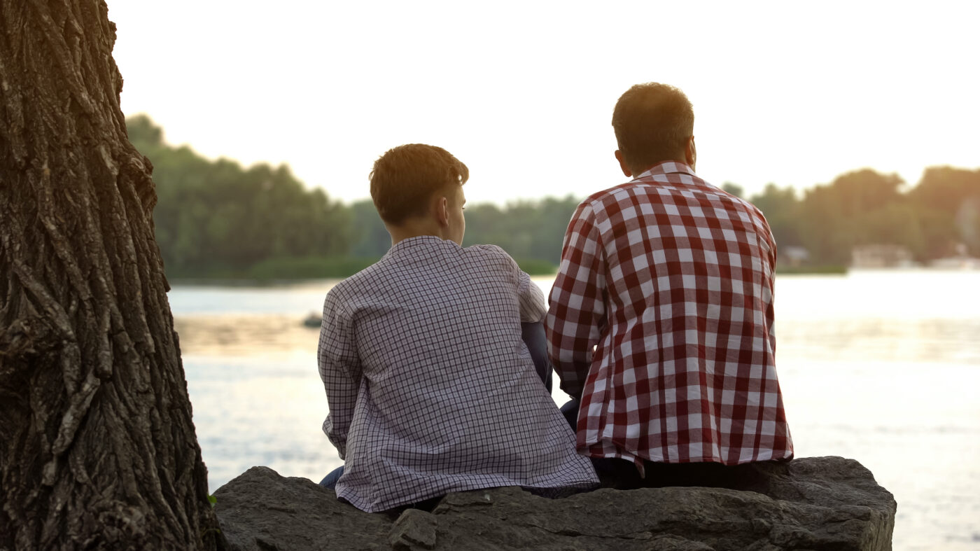 Male teenager and his father sitting on stone near lake and talking about life